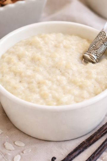 French rice pudding (riz au lait) in a white ramekin with vanilla beans and cinnamon sticks, styled on a neutral linen background