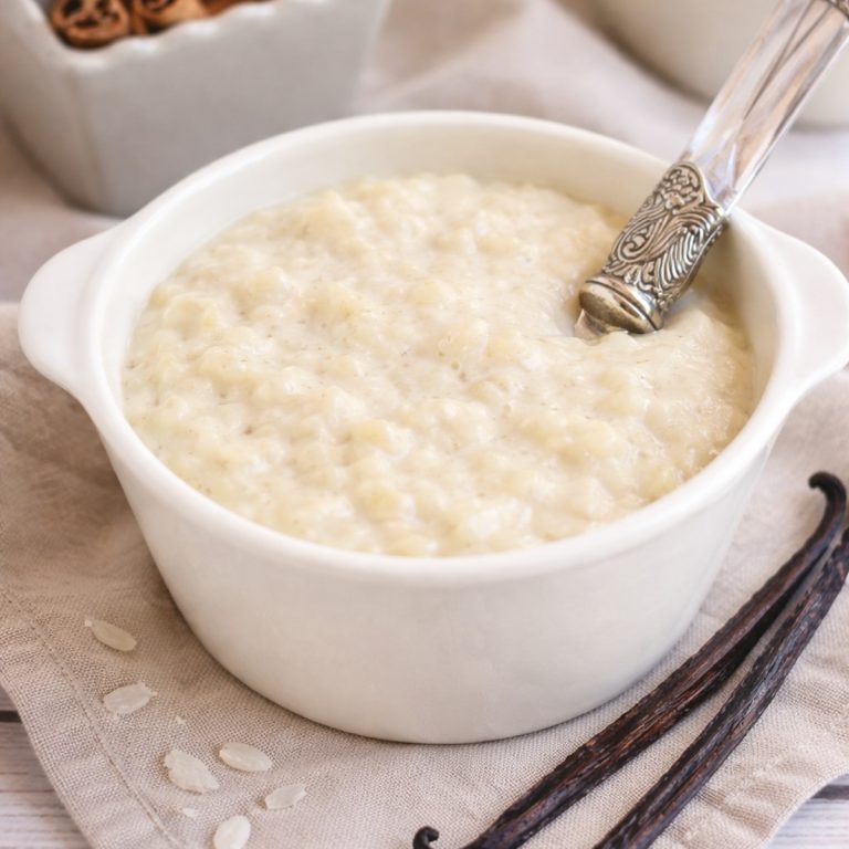 French rice pudding (riz au lait) in a white ramekin with vanilla beans and cinnamon sticks, styled on a neutral linen background