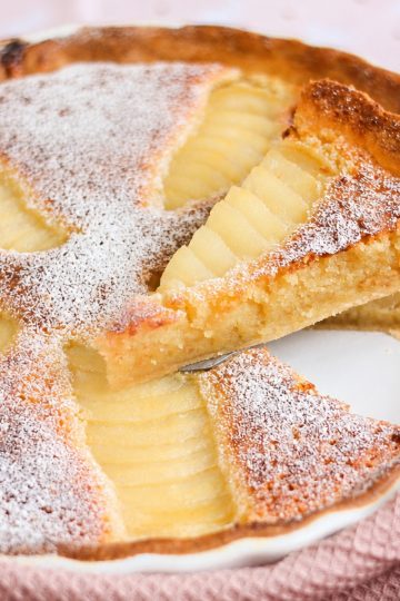 Homemade pear frangipane tart on a pink cloth with one slice lifted to show the almond filling.