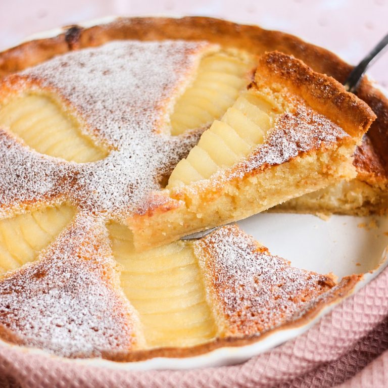 Homemade pear frangipane tart on a pink cloth with one slice lifted to show the almond filling.