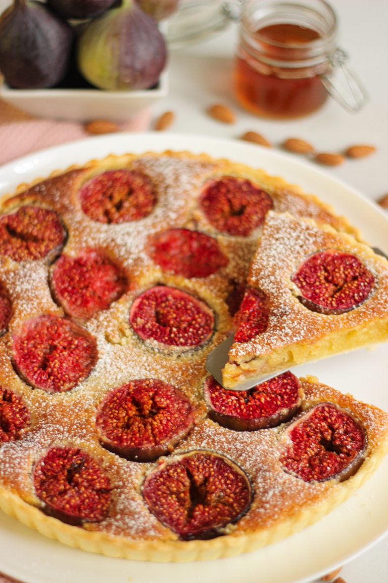 Fig tart dusted with powdered sugar on a white plate, with one slice cut and lifted on a pie server.