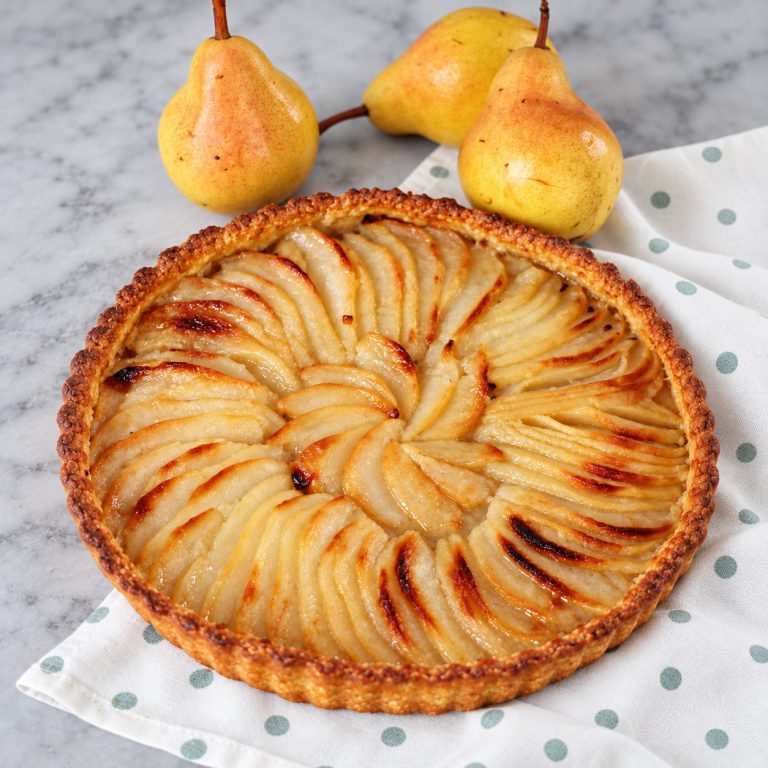 French pear tart with overlapping golden pear slices and a buttery crust, with fresh pears behind it.
